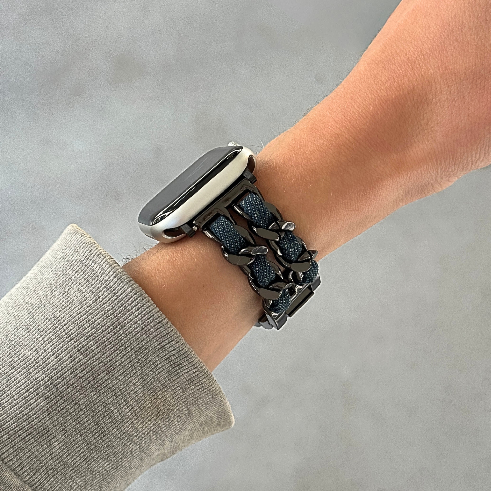 A close-up of a person's wrist wearing a black metallic chain link Apple watch band with a textured denim fabric inner lining, paired with a smartwatch.
