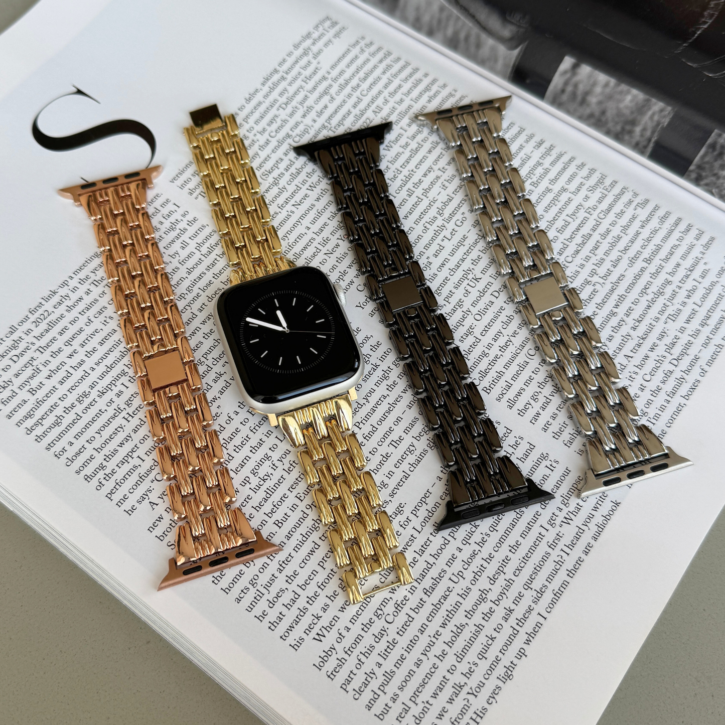 Rose gold, gold, black and silver link Apple Watch bands laid out on a magazine. Sleek metal iWatch straps and armbands, compatible with Apple Watch 38mm to 49mm, Series 1-11, SE and Ultra