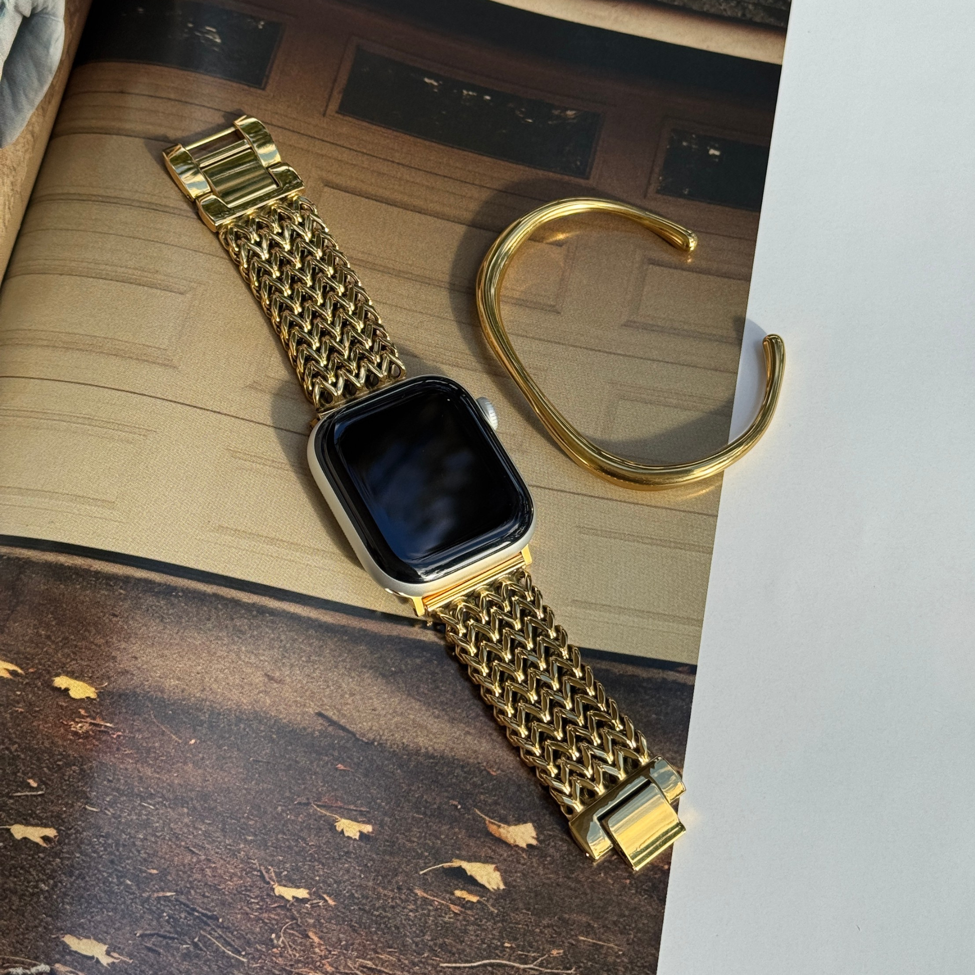 A gold tone metal mesh watch band attached to an Apple smartwatch, displayed diagonally on a textured surface next to a matching gold cuff bracelet.