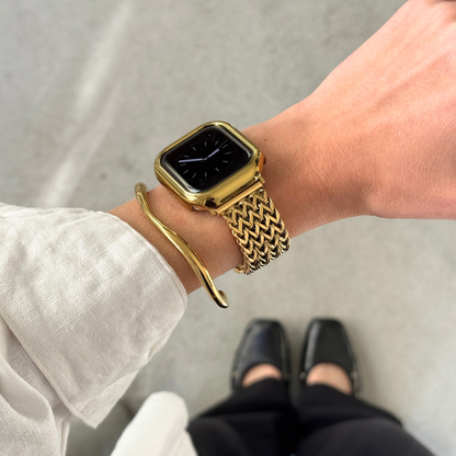 A close-up of a person's wrist wearing a gold toned Apple smartwatch with a woven mesh band, along with a slim gold bangle bracelet and a gold matching bezel. The photo is taken from above, showing the person's arm, hand, sleeve, pants, and dark shoes against concrete floor.