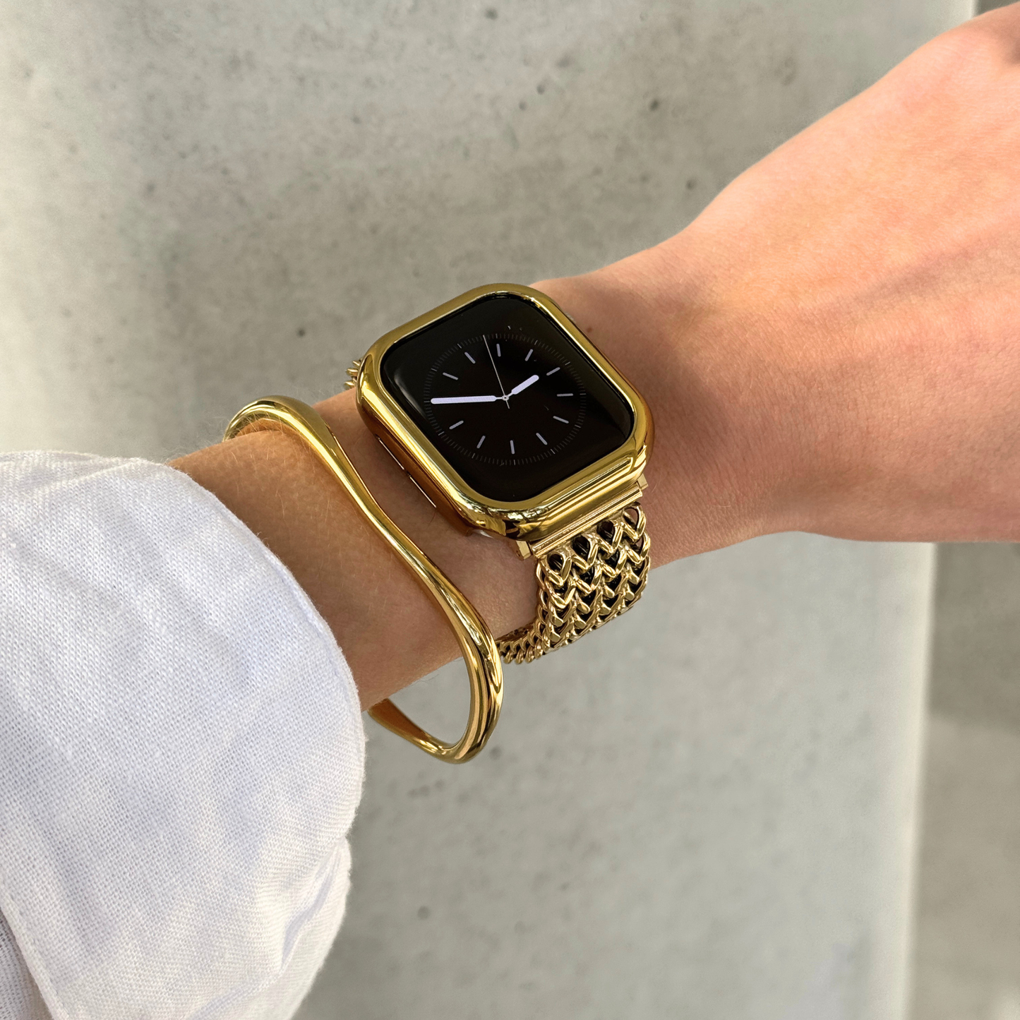 A close-up of a person's wrist wearing a gold toned Apple watch with a textured gold link band, along with a matching gold bangle bracelet and a matching watch case cover.