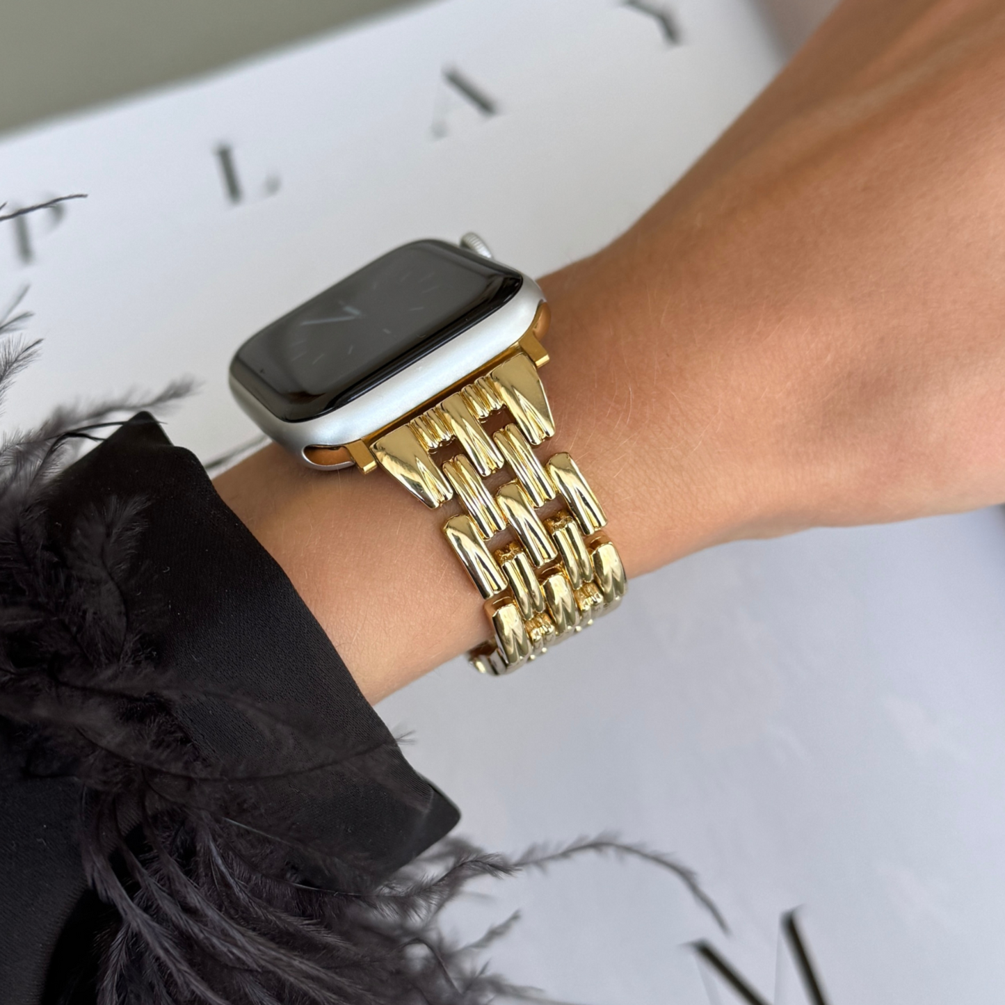 Gold link Apple Watch band worn on a wrist with a feather trimmed cuff. Fashion forward metal iWatch bracelet, compatible with Apple Watch 38mm to 49mm, Series 1-11, SE and Ultra