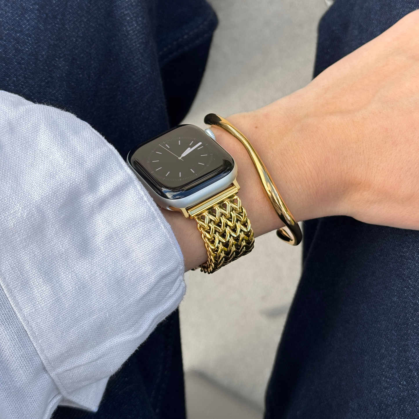 A close-up of a person's wrist wearing a gold mesh metal watch band on an Apple smartwatch, along with a gold bangle bracelet, a linen shirt cuff, and dark jeans.
