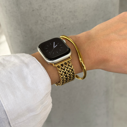 A close-up of a person's wrist wearing a gold mesh watch band on an Apple Watch, paired with a gold bangle bracelet, against a concrete wall background.