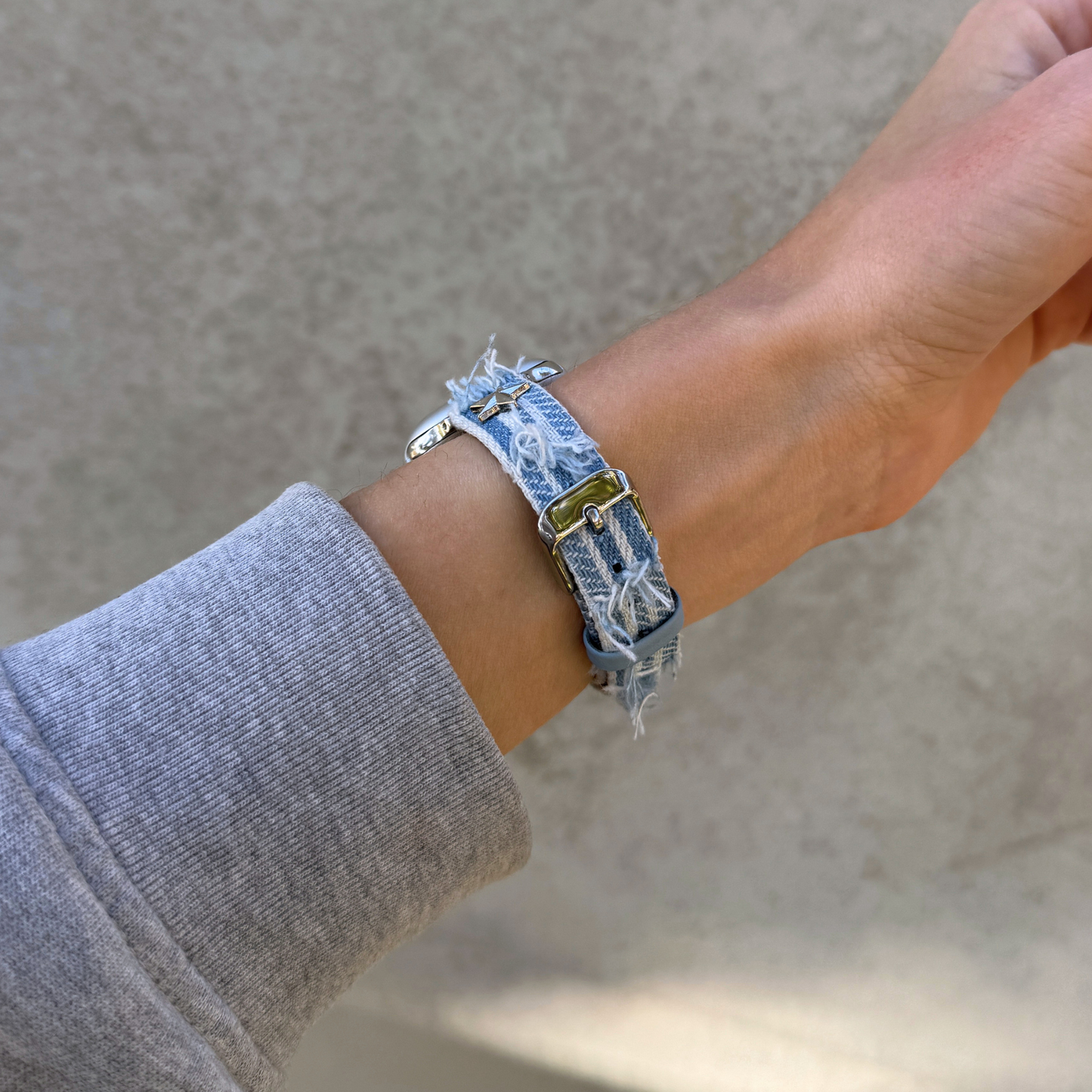 A close-up of a person's wrist wearing a light blue denim Apple Watch bracelet with distressed frayed edges and a silver-toned buckle, paired with a gray long-sleeve shirt against a neutral concrete background.