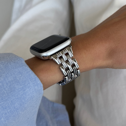 Silver link Apple Watch band worn on a wrist with a blue shirt cuff. Feminine metal iWatch strap that elevates any outfit, compatible with Apple Watch 38mm to 49mm, Series 1-11, SE and Ultra