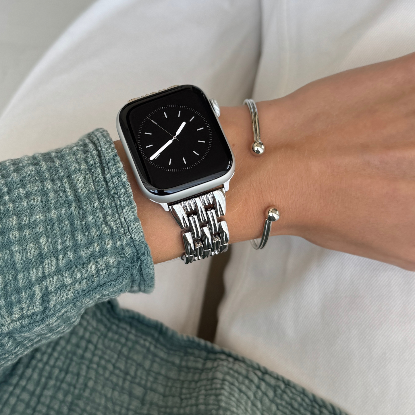 Silver link Apple Watch band worn with a green textured sleeve and a silver bangle. Feminine metal iWatch bracelet, compatible with Apple Watch 38mm to 49mm, Series 1-11, SE and Ultra