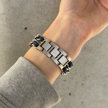 A close-up of a person's wrist wearing a starlight toned metal link Apple Watch bracelet with dark denim braided cord accents, paired with a long-sleeve shirt against a concrete background.