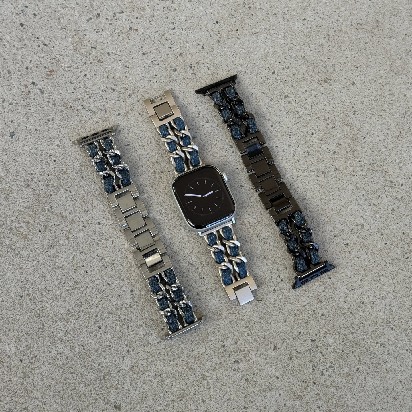 Three metal watch bands arranged on a light gray concrete surface, on an Apple watch. The left band is silver with dark blue denim accents, the center band is starlight with dark blue denim accents, and the right band is black with dark blue denim accents.
