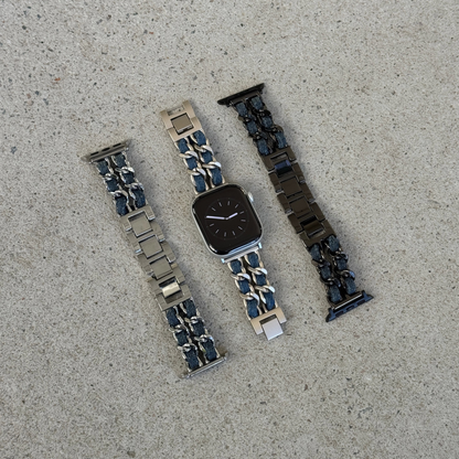 Three metal watch bands arranged on a light gray concrete surface, on an Apple watch. The left band is silver with dark blue denim accents, the center band is starlight with dark blue denim accents, and the right band is black with dark blue denim accents.