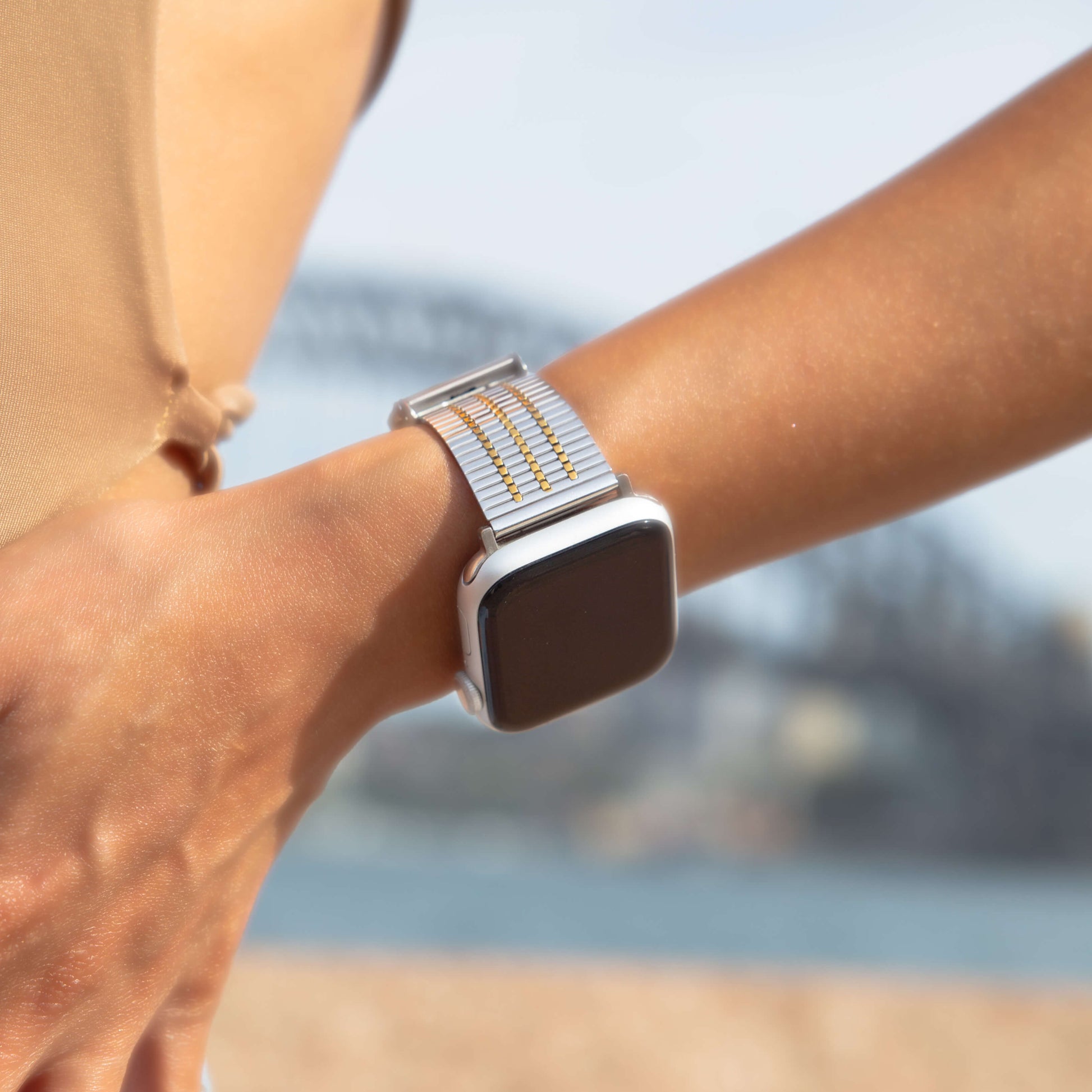 Silver gold flat woven Apple Watch band worn on a wrist beside the water. Waterproof stainless steel iWatch bracelet, compatible with Apple Watch 38mm to 49mm, Series 1-11, SE and Ultra