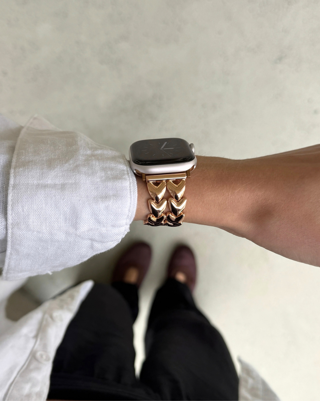 A close-up of a person's wrist wearing a rose-gold-toned metal link Apple watch band, shown from a top-down perspective against a light gray floor.