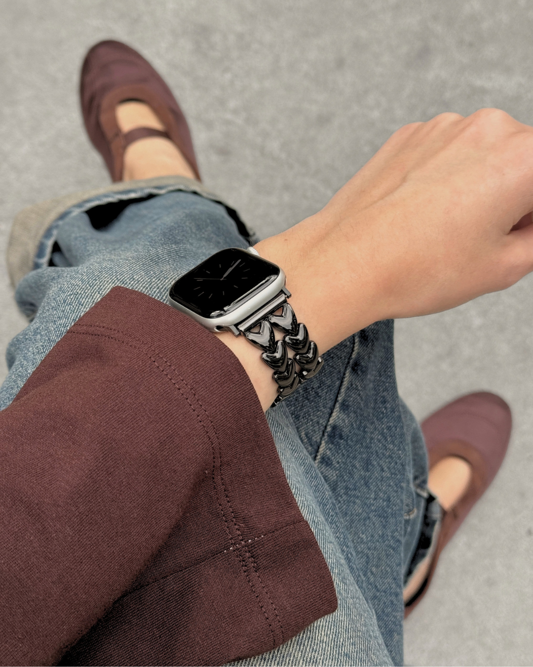 A close-up lifestyle shot of a person wearing a chunky, textured black metallic heart-shape link watch band on an Apple Watch, paired with rolled-up blue jeans, a brown blazer, and brown Mary Jane flats on a gray concrete background.