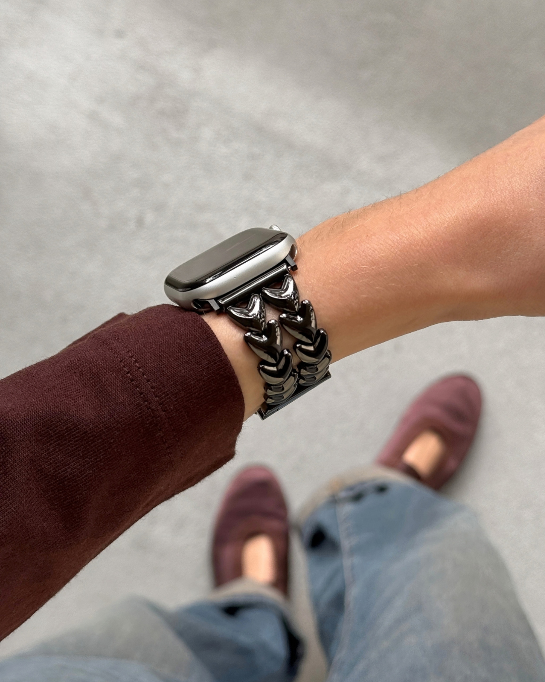 A close-up of a person's wrist wearing a chunky, black metal heart-shape link watch band with an Apple Watch. The person is wearing a dark burgundy long-sleeve top, light blue jeans, and burgundy slip-on shoes, standing on a gray concrete floor.