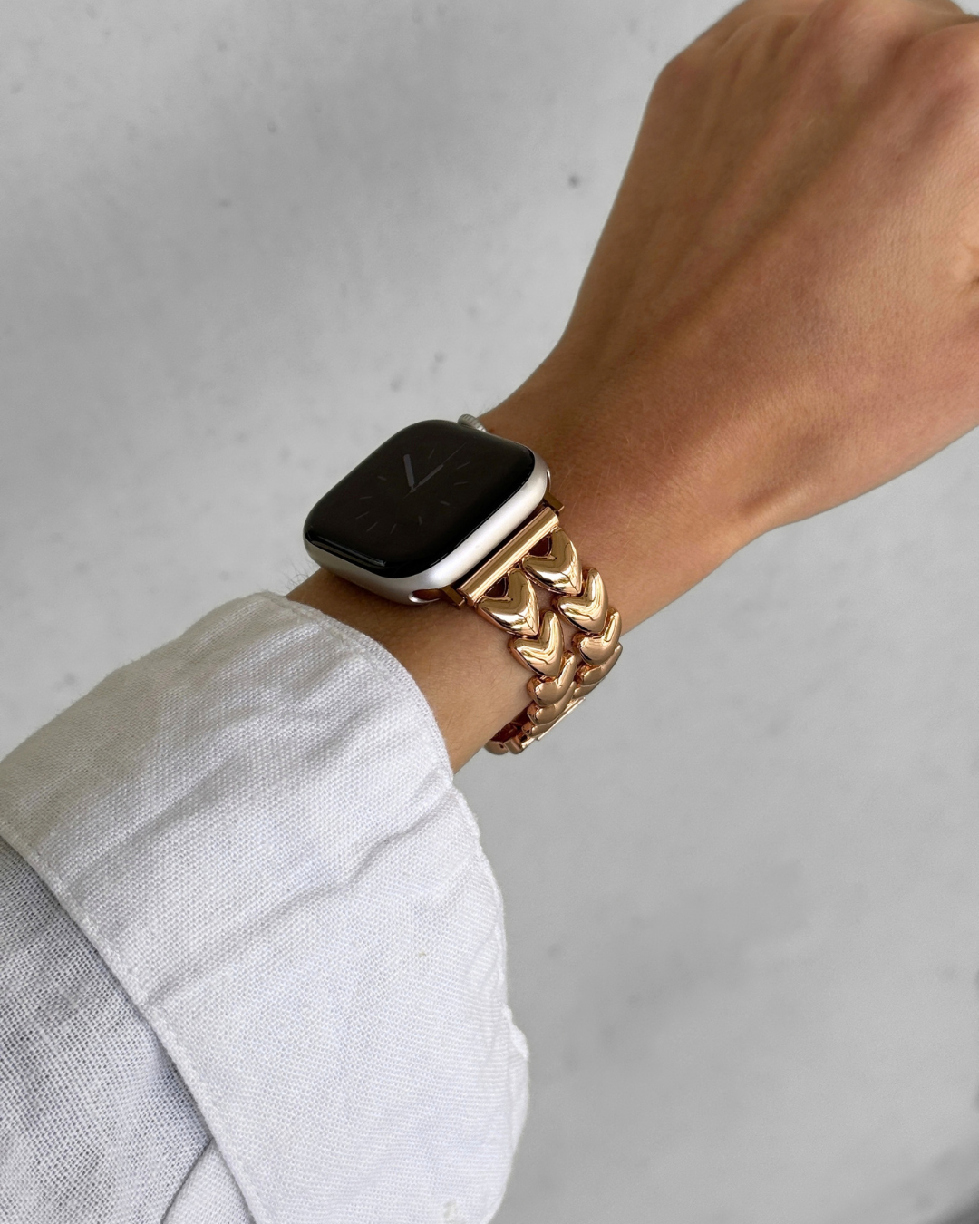 A close-up of a person's wrist wearing a rose gold-toned metal Apple watch band, set against a light gray textured background.
