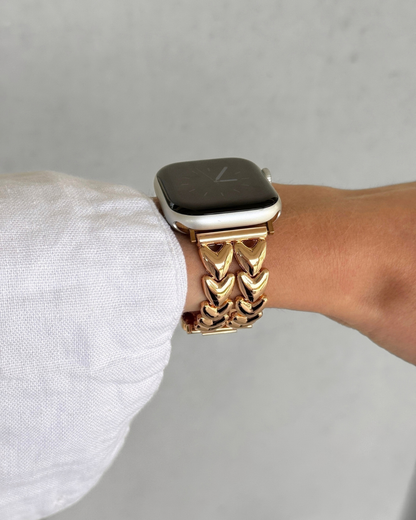 A close-up of a rose-gold heart link chain watch band worn on an Apple Watch, shown on a person's wrist with a white sleeve visible.