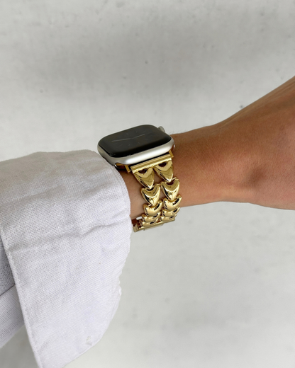 A close-up of a person's wrist wearing a gold-toned heart-shaped link watch band with an Apple Watch, set against a light gray textured background.