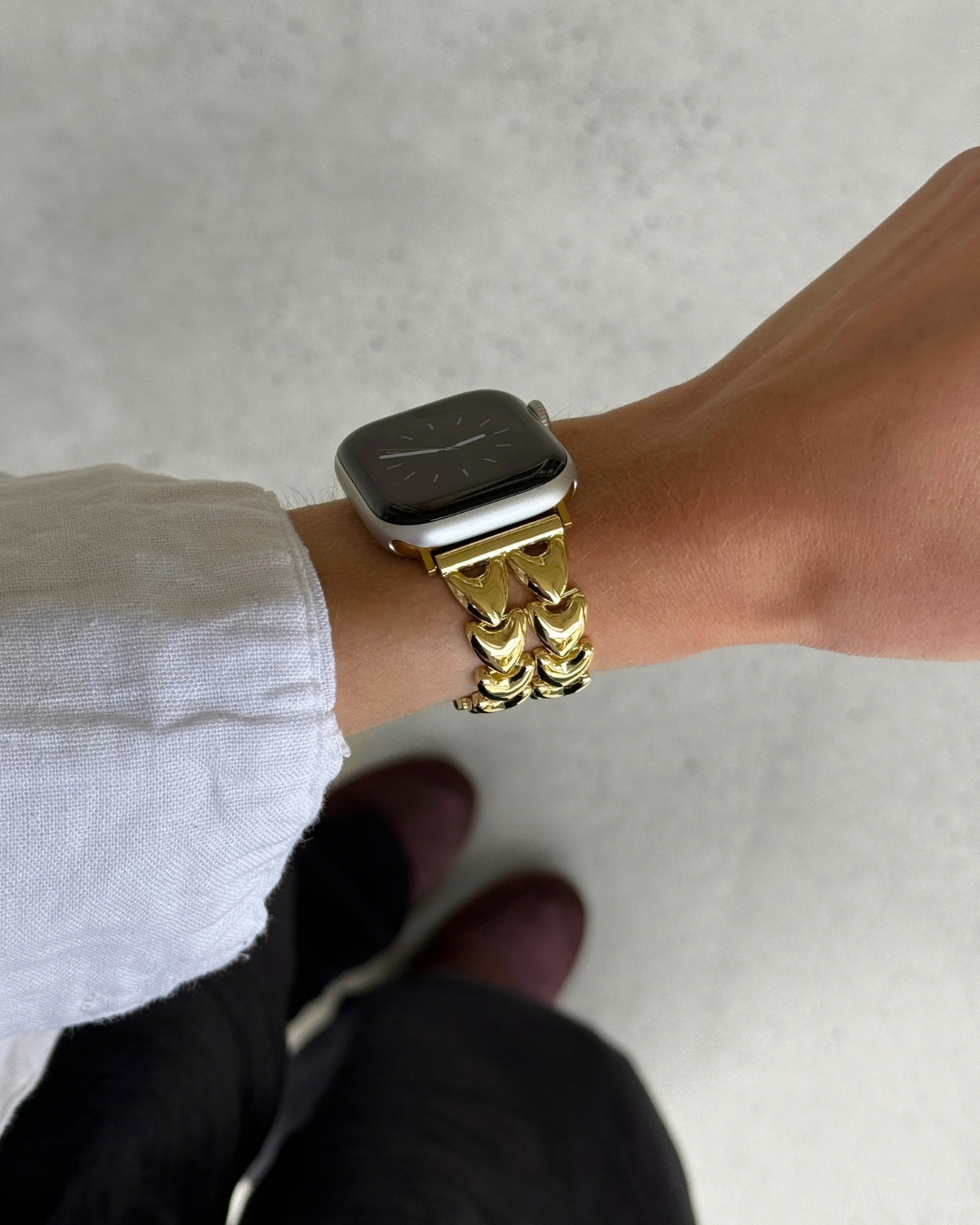 A close-up of a person's wrist wearing a chunky, heart-shaped gold chain watch strap on an Apple Watch, with a white shirt cuff and dark pants visible against a light gray concrete floor.