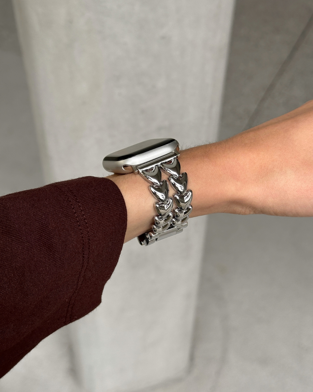 A close-up of a person's wrist wearing a chunky, polished heart-shaped silver metal link watch band with an Apple Watch, set against a neutral gray background.