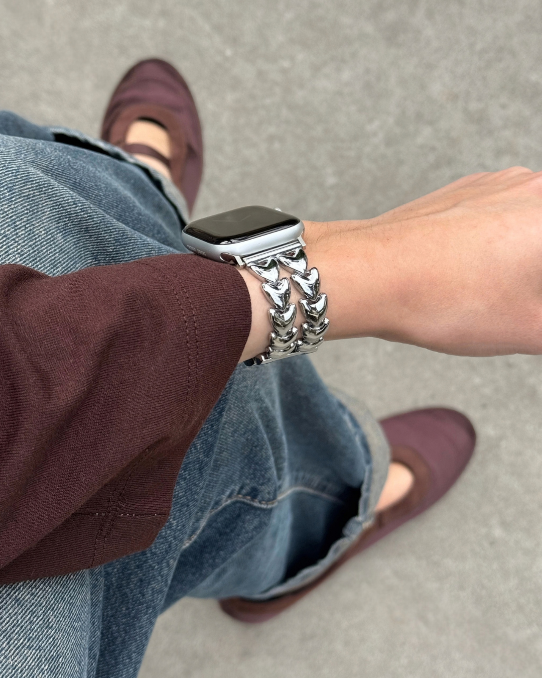 A close-up lifestyle shot of a person wearing a chunky silver heart-shaped chain link watch band on an Apple Watch. The person is wearing blue jeans, a dark burgundy top, and burgundy flat shoes, standing on a gray concrete floor.