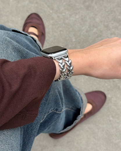 A close-up lifestyle shot of a person wearing a chunky silver heart-shaped chain link watch band on an Apple Watch. The person is wearing blue jeans, a dark burgundy top, and burgundy flat shoes, standing on a gray concrete floor.