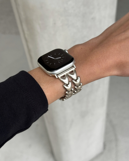 A close-up of a person's wrist wearing a starlight colour heart-shaped metal link watch band on an Apple Watch, against a light gray concrete background.