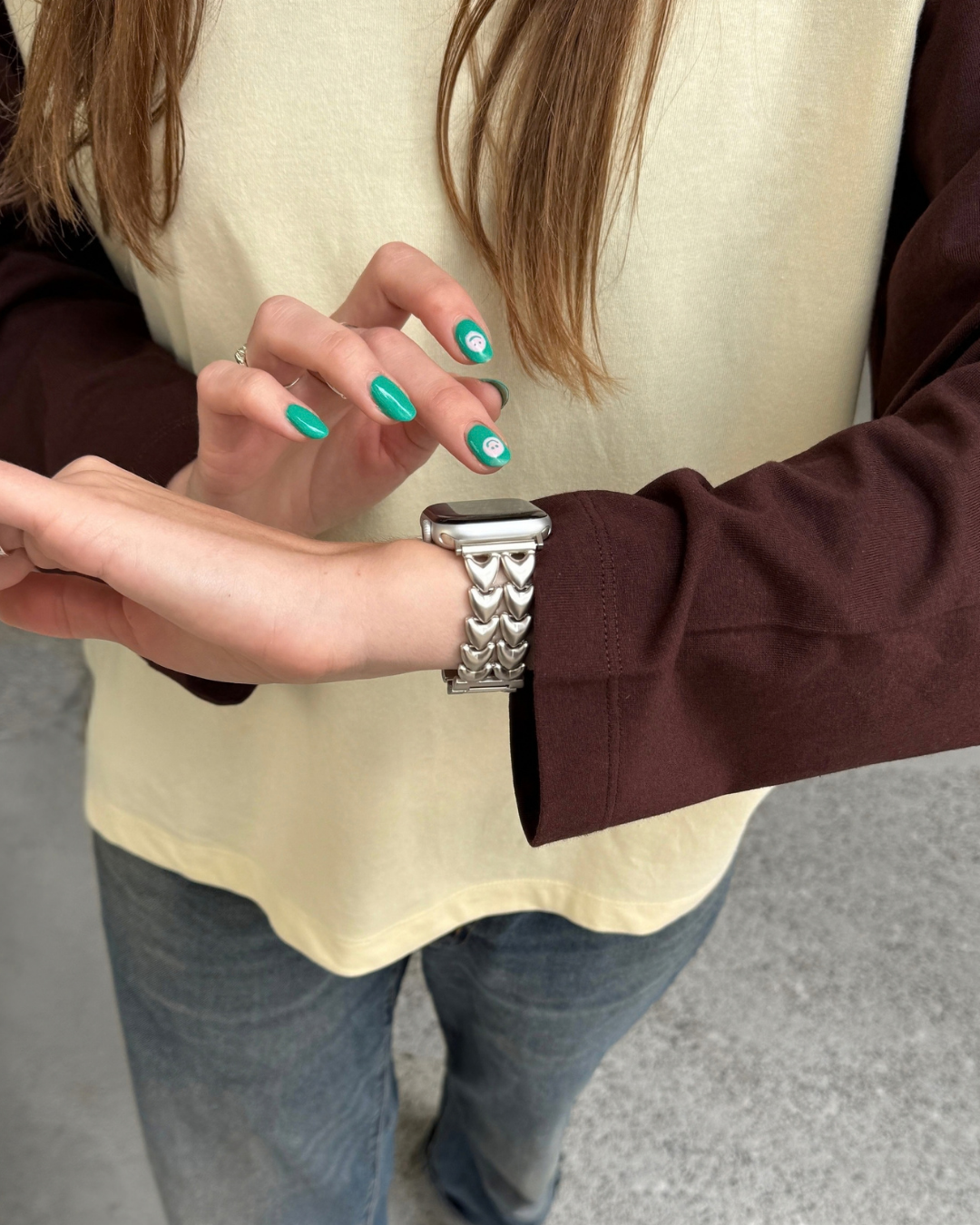 A close-up of a person wearing a starlight Apple Watch band with a heart-shaped metal links, paired with a yellow top, brown sleeves, and blue jeans.