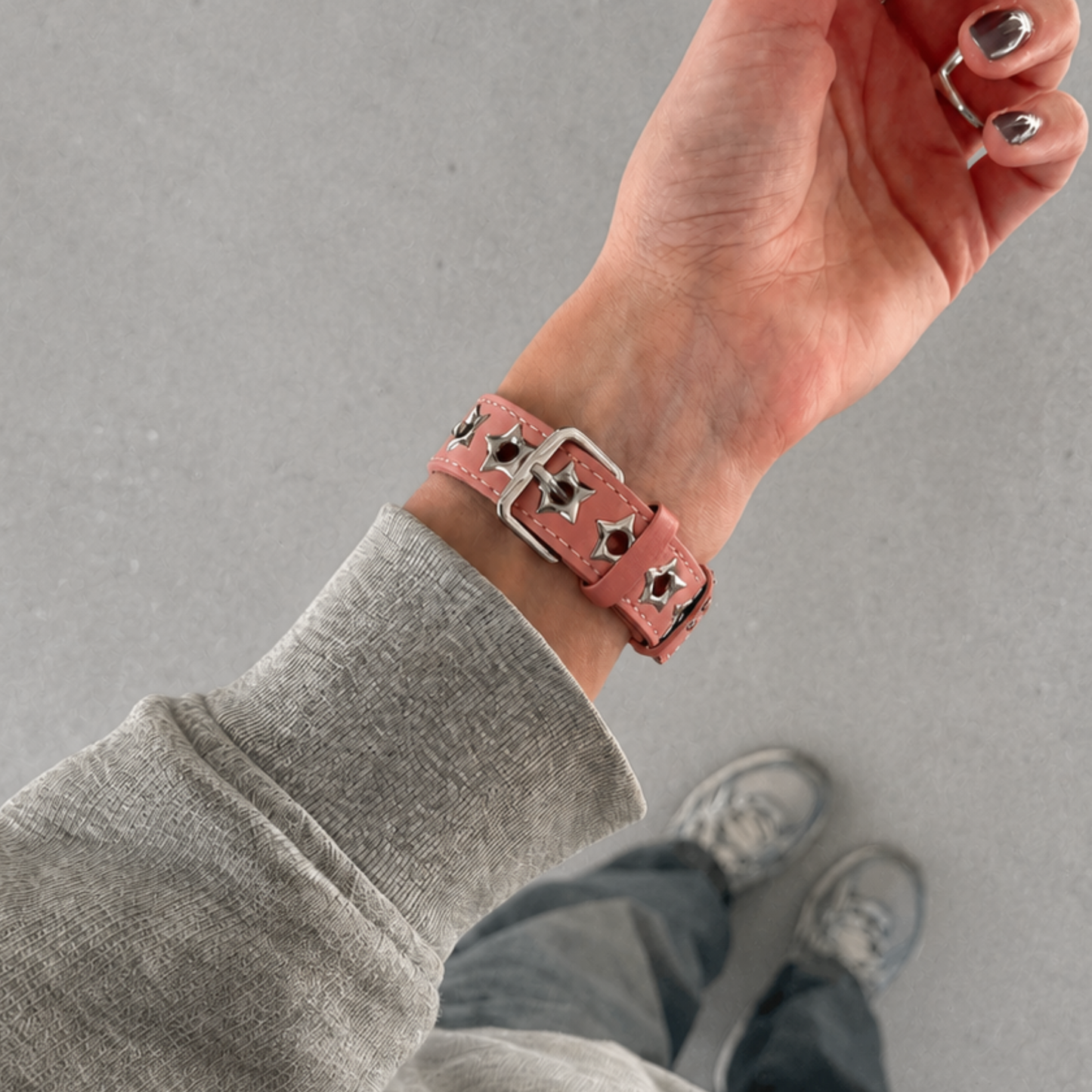 A close-up of a person's wrist wearing a pink faux-leather studded belt-style Apple watch band with silver-toned hardware, shown against a gray pavement background.