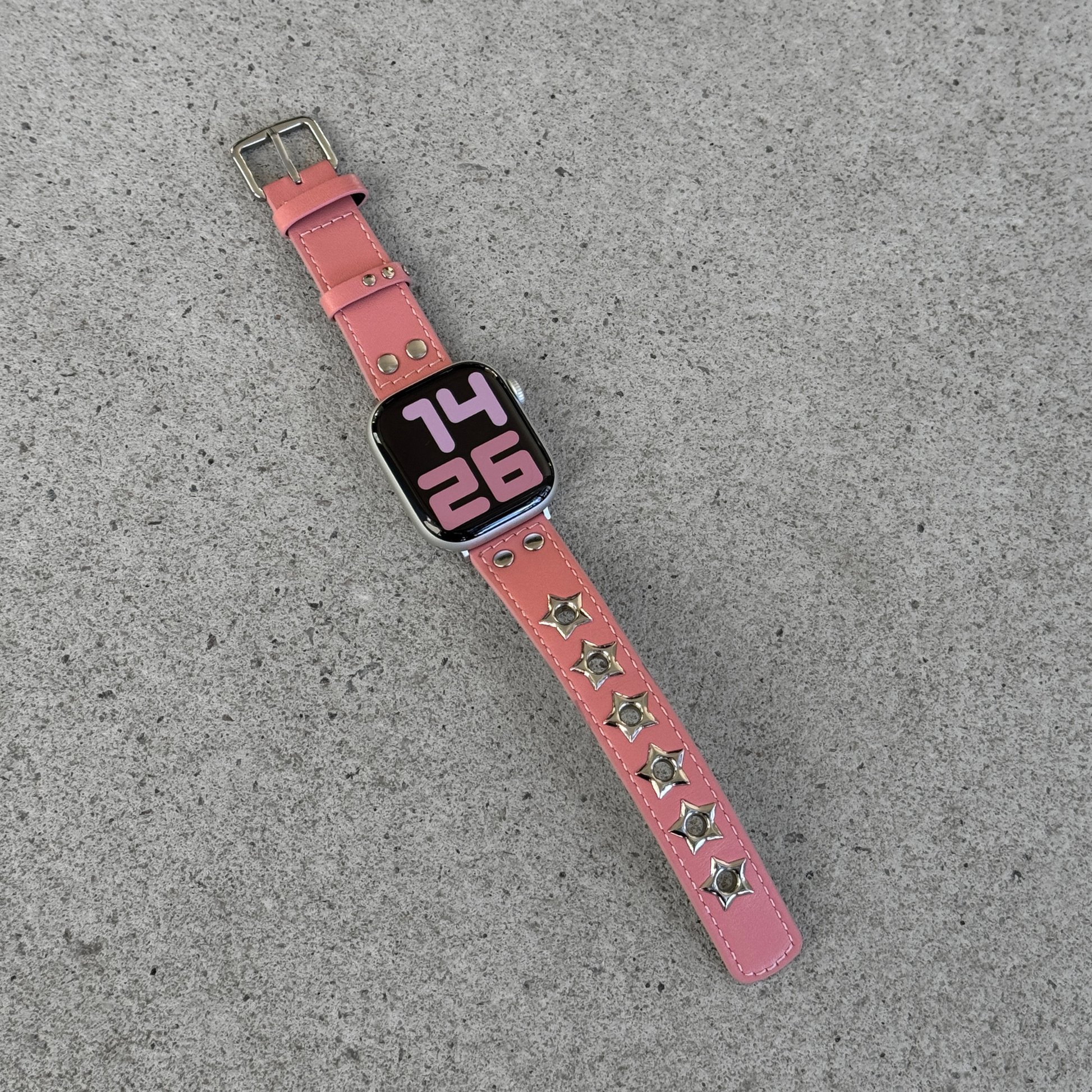 A pink leather watch band with silver star-shaped studs, shown attached to an Apple Watch on a textured gray concrete surface.