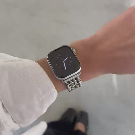 A close-up video of a person's wrist wearing a silver metal mesh watch band with an Apple Watch. The person is wearing a white long-sleeve shirt and black pants, standing on a light gray concrete floor.