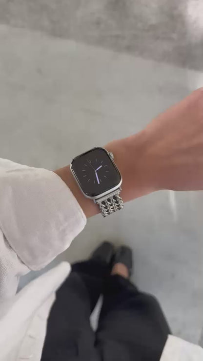 A close-up video of a person's wrist wearing a silver metal mesh watch band with an Apple Watch. The person is wearing a white long-sleeve shirt and black pants, standing on a light gray concrete floor.