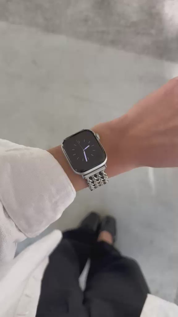 A close-up video of a person's wrist wearing a silver metal mesh watch band with an Apple Watch. The person is wearing a white long-sleeve shirt and black pants, standing on a light gray concrete floor.