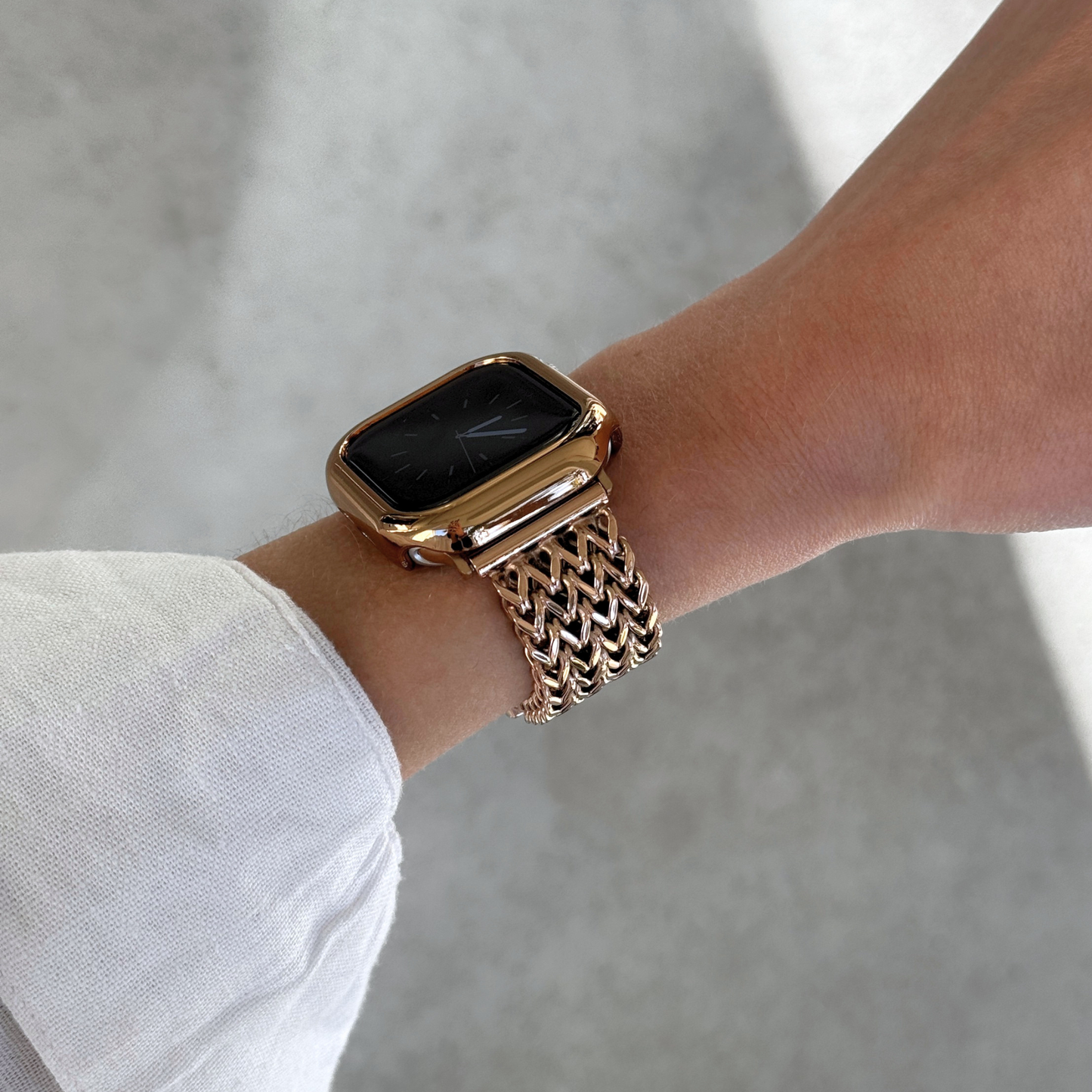 A close-up of a person's wrist wearing a rose gold toned Apple smartwatch with a matching metallic mesh band and bezel, set against a soft concrete background.