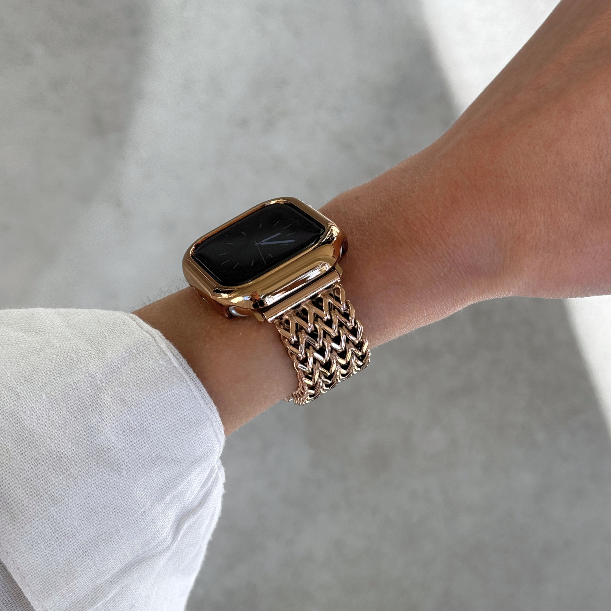 A close-up of a person's wrist wearing a rose gold toned Apple smartwatch with a matching metallic mesh band and bezel, set against a soft concrete background.