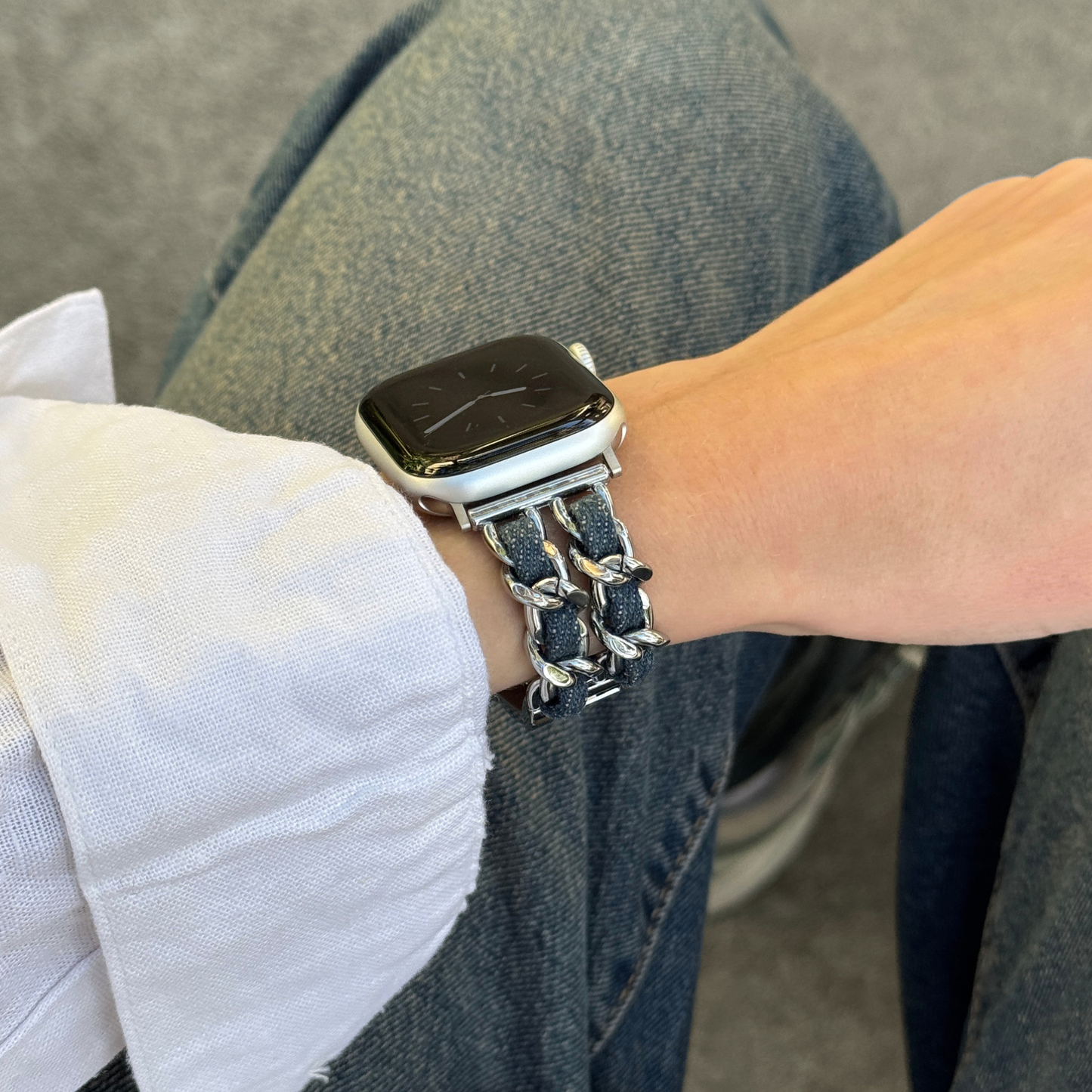 A close-up of a person's wrist wearing a silver toned metal Apple watch band with dark woven accents, paired with a smartwatch, jeans, and a shirt.