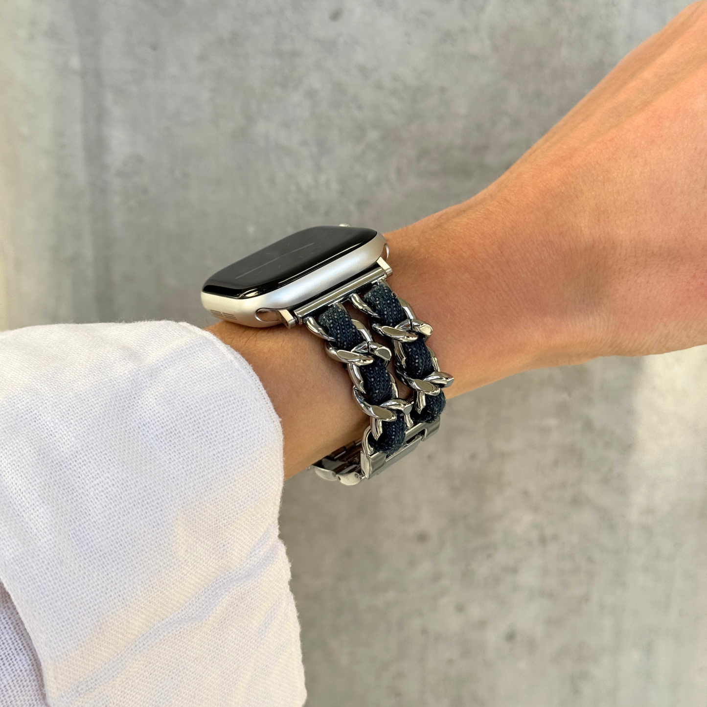 A close-up of a person's wrist wearing a silver toned metal link bracelet watch band with dark denim woven fabric details, attached to an Apple Watch.