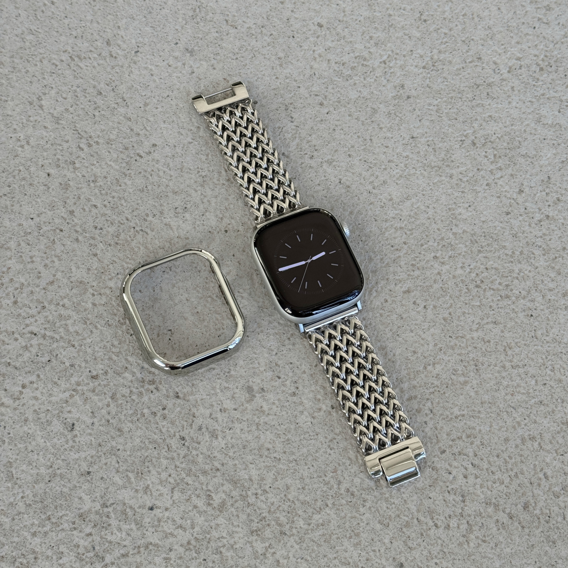 A silver toned metal mesh watch band with a matching bezel protector, shown laid flat on a textured surface next to an apple smartwatch.