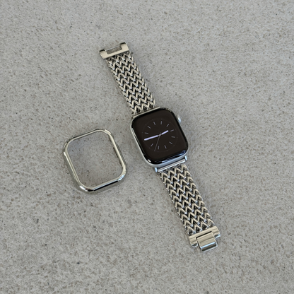 A silver toned metal mesh watch band with a matching bezel protector, shown laid flat on a textured surface next to an apple smartwatch.