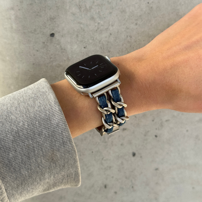 A close-up of a person's wrist wearing an Apple smartwatch with a braided denim and starlight coloured chain strap, set against concrete background.