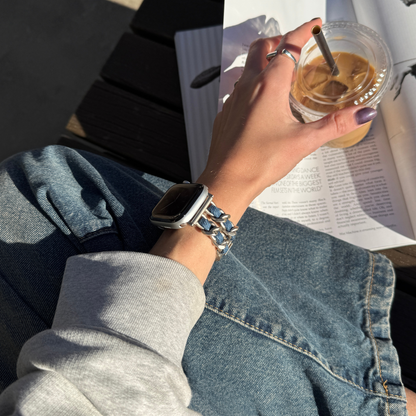 A close-up lifestyle shot of a person wearing a starlight chain-link Apple smartwatch band on their wrist, sitting outdoors next to an open book and holding an iced coffee.
