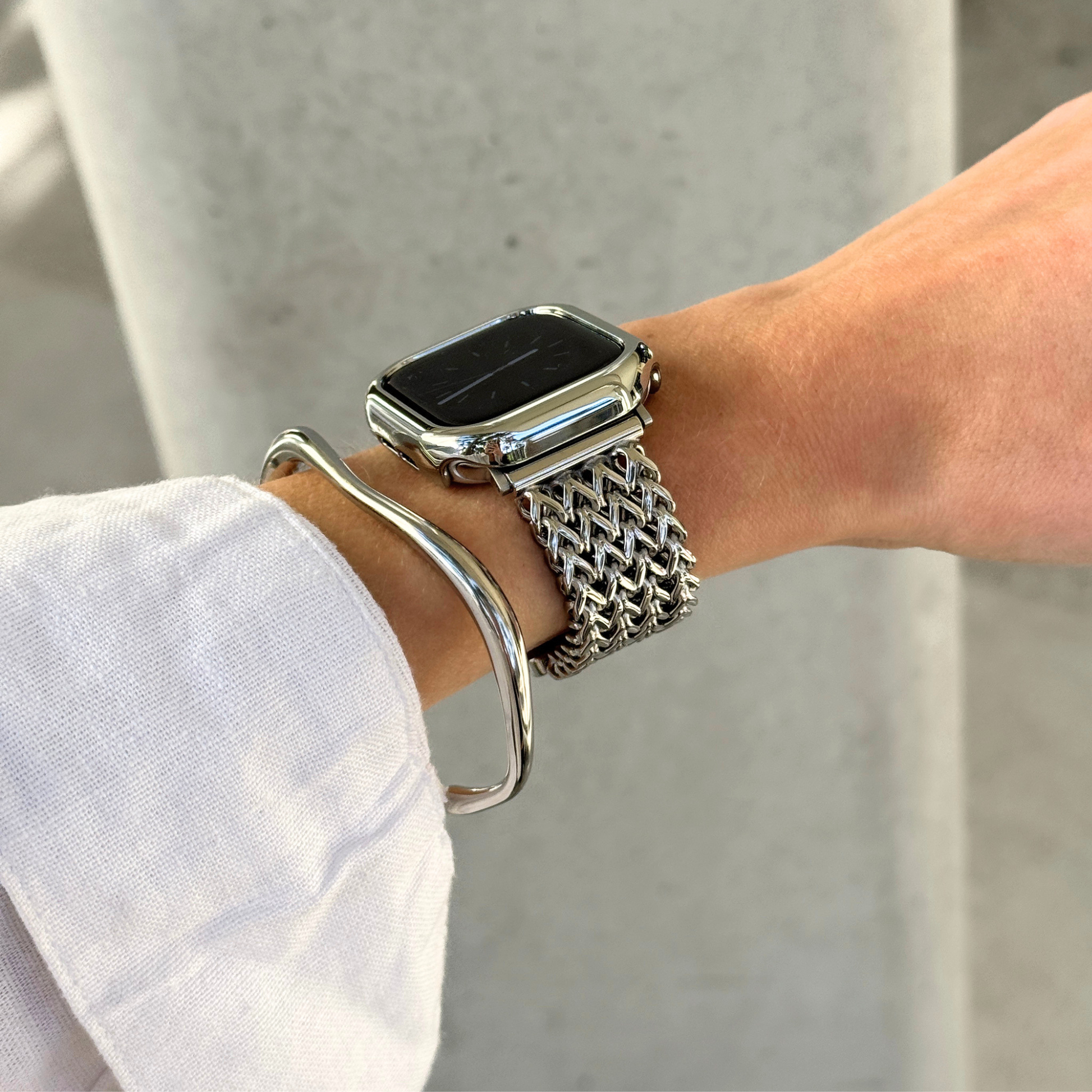 A close-up of a person's wrist wearing a silver mesh Apple watch band with a smartwatch, along with a silver bangle bracelet and a matching bezel, against a light concrete background.