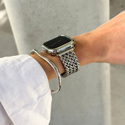 A close-up of a person's wrist wearing a silver mesh Apple watch band with a smartwatch, along with a silver bangle bracelet and a matching bezel, against a light concrete background.