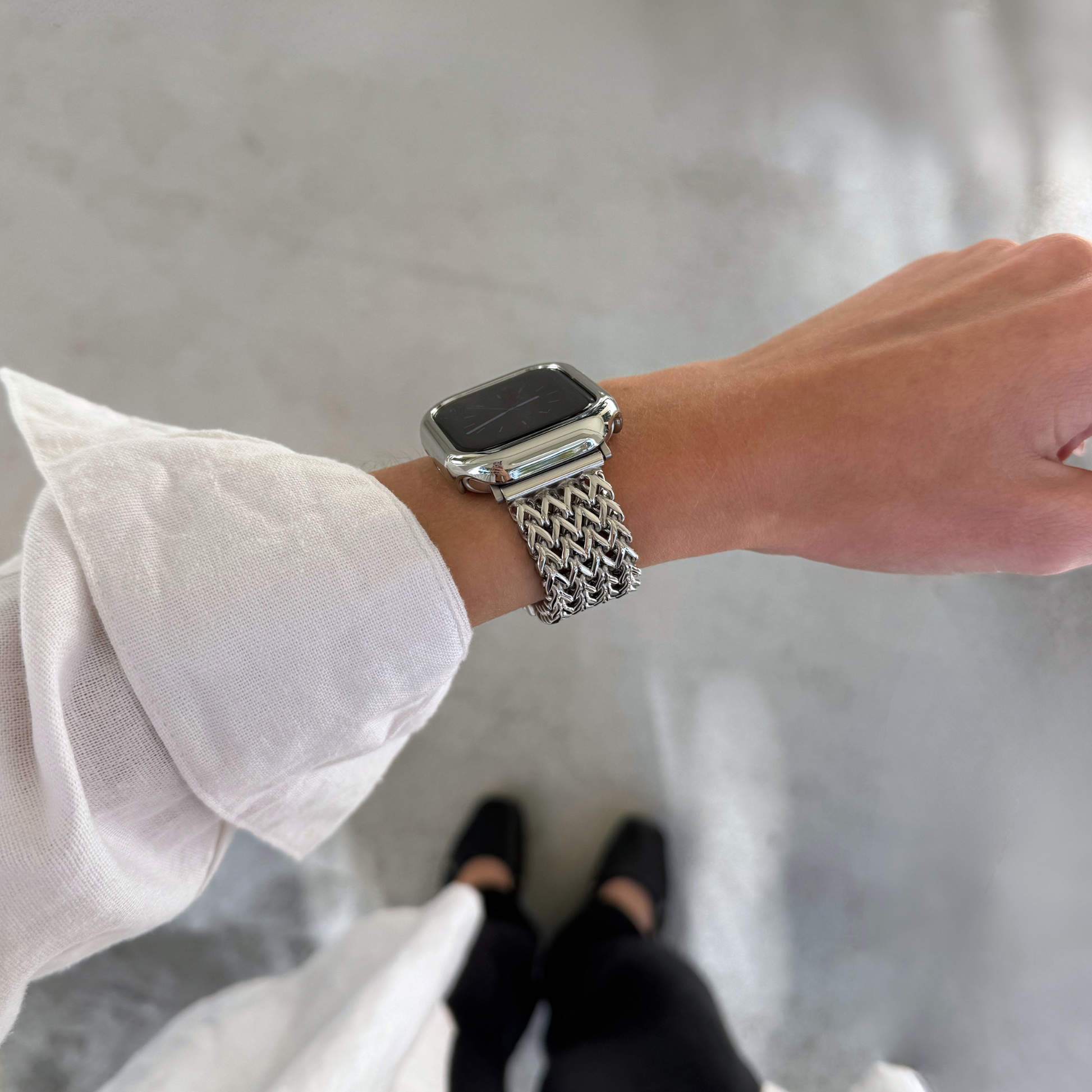 A close-up of a person's wrist wearing a silver metallic mesh Apple Watch band and a matching bezel cover, with a linen shirt sleeve visible and concrete floor in the background.