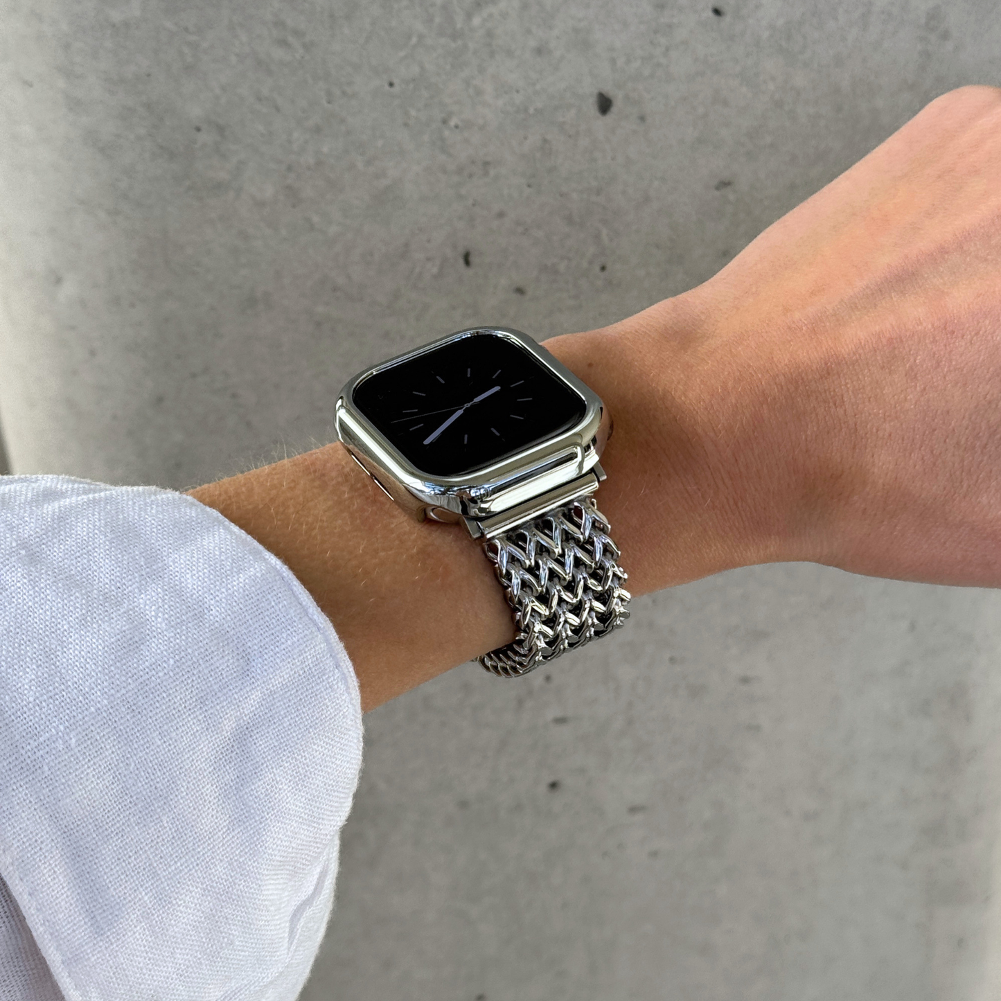 A close-up of a person's wrist wearing a silver smartwatch with a shiny metallic mesh Apple watch band and a matching case cover, set against a concrete wall.