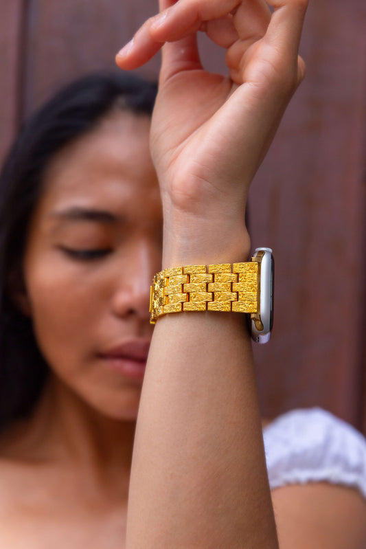 Gold baroque pattern Apple Watch band worn on a raised wrist against a wooden wall. Vintage inspired metal iWatch bracelet with a luxurious look, compatible with Apple Watch 38mm, 40mm, 41mm, 42mm, 44mm, 45mm, 46mm and 49mm, Series 1-11, SE and Ultra