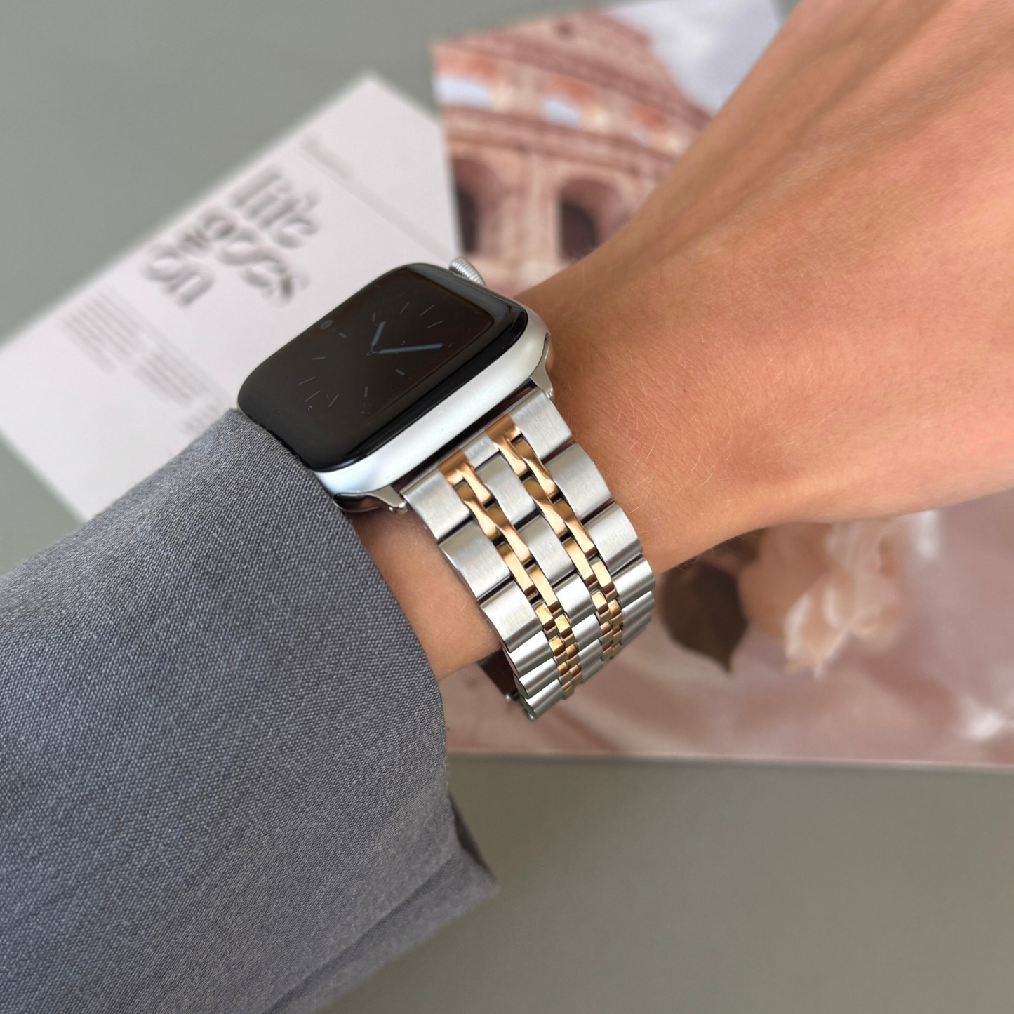 Silver rose gold link Apple Watch band worn on a wrist beside a travel magazine. Waterproof stainless steel iWatch bracelet, compatible with Apple Watch 38mm to 49mm, Series 1-11, SE and Ultra