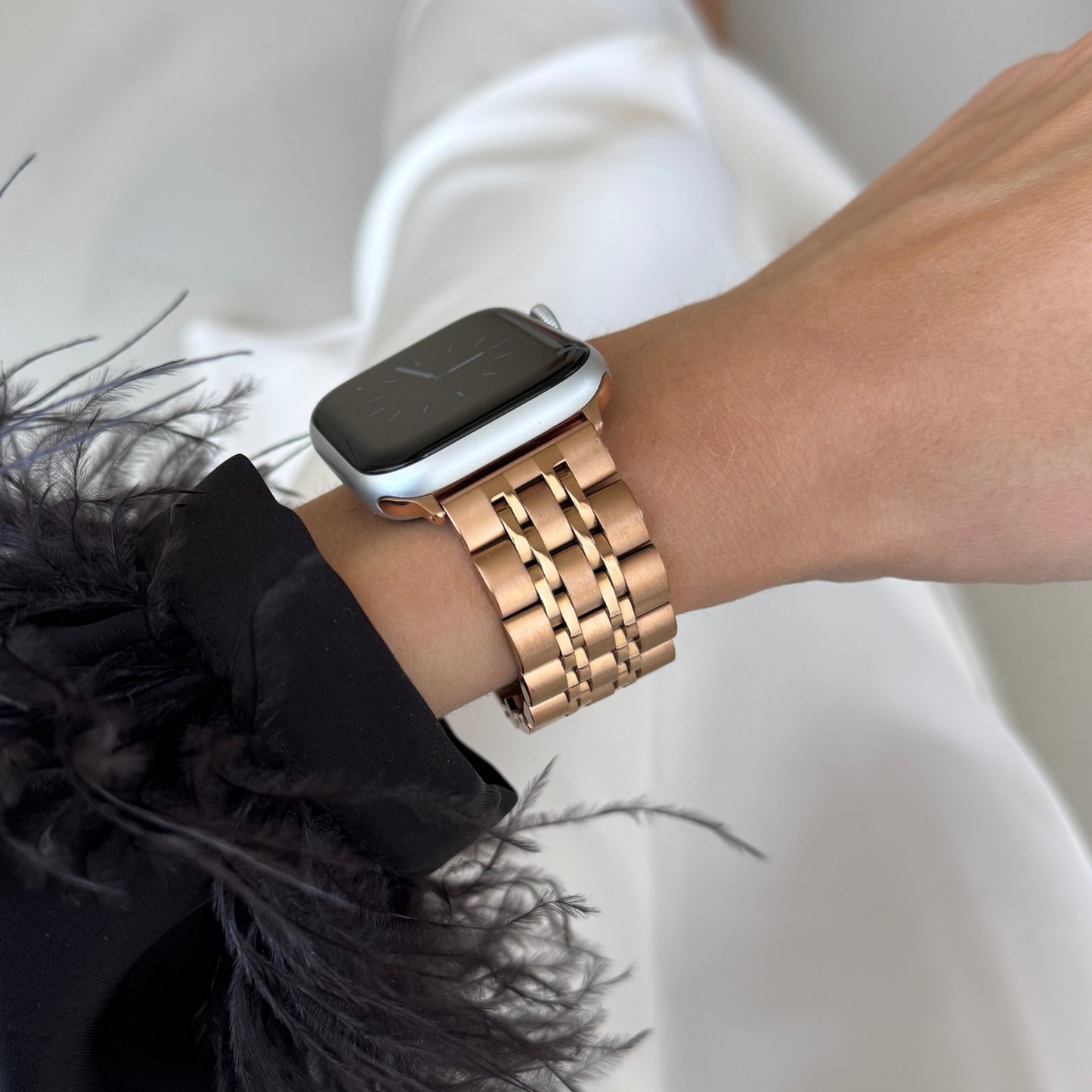 Rose gold link Apple Watch band worn on a wrist with a feather trimmed cuff. Waterproof stainless steel iWatch bracelet, compatible with Apple Watch 38mm, 40mm, 41mm, 42mm, 44mm, 45mm, 46mm and 49mm, Series 1-11, SE and Ultra