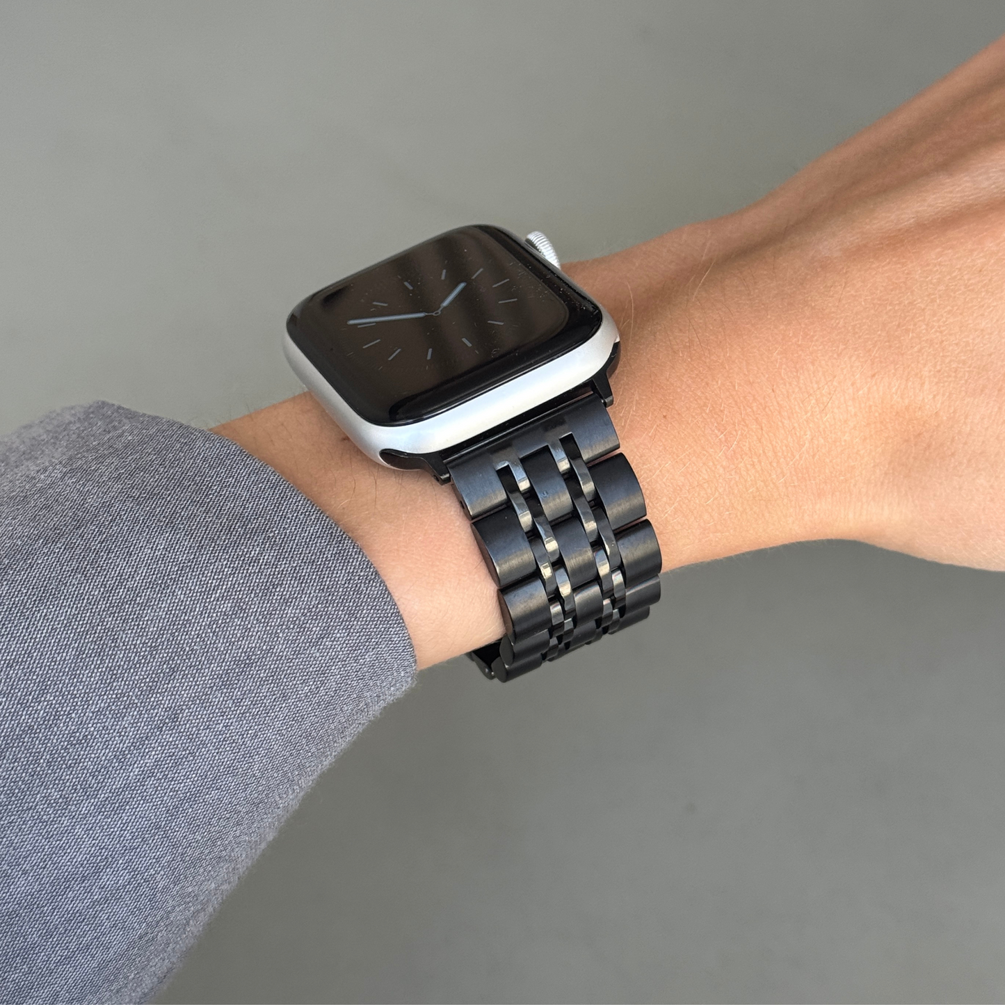 Black link Apple Watch band worn on a wrist with a grey sleeve. Minimalist stainless steel iWatch strap, compatible with Apple Watch 38mm to 49mm, Series 1-11, SE and Ultra