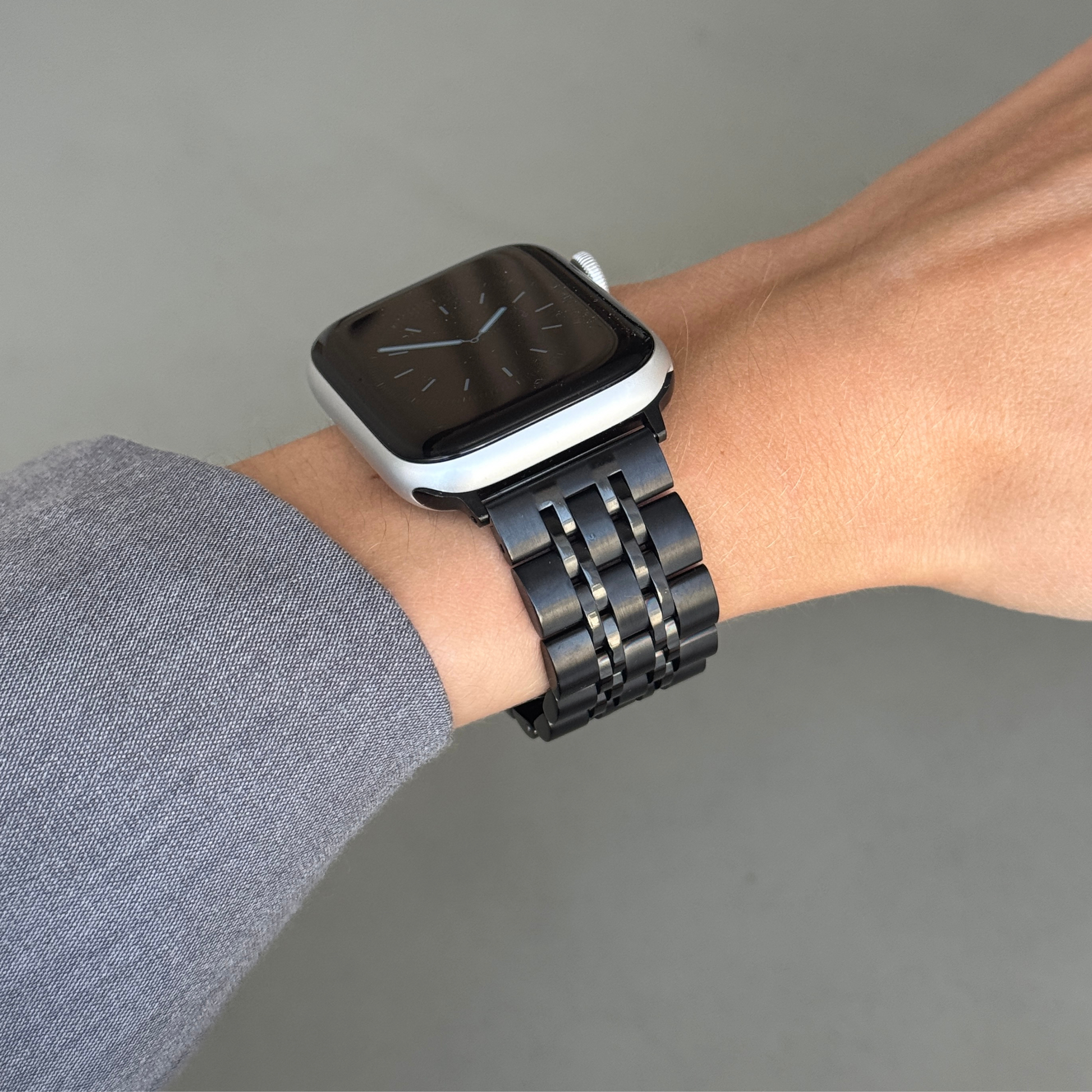 Black link Apple Watch band worn on a wrist with a grey sleeve. Minimalist stainless steel iWatch strap, compatible with Apple Watch 38mm to 49mm, Series 1-11, SE and Ultra