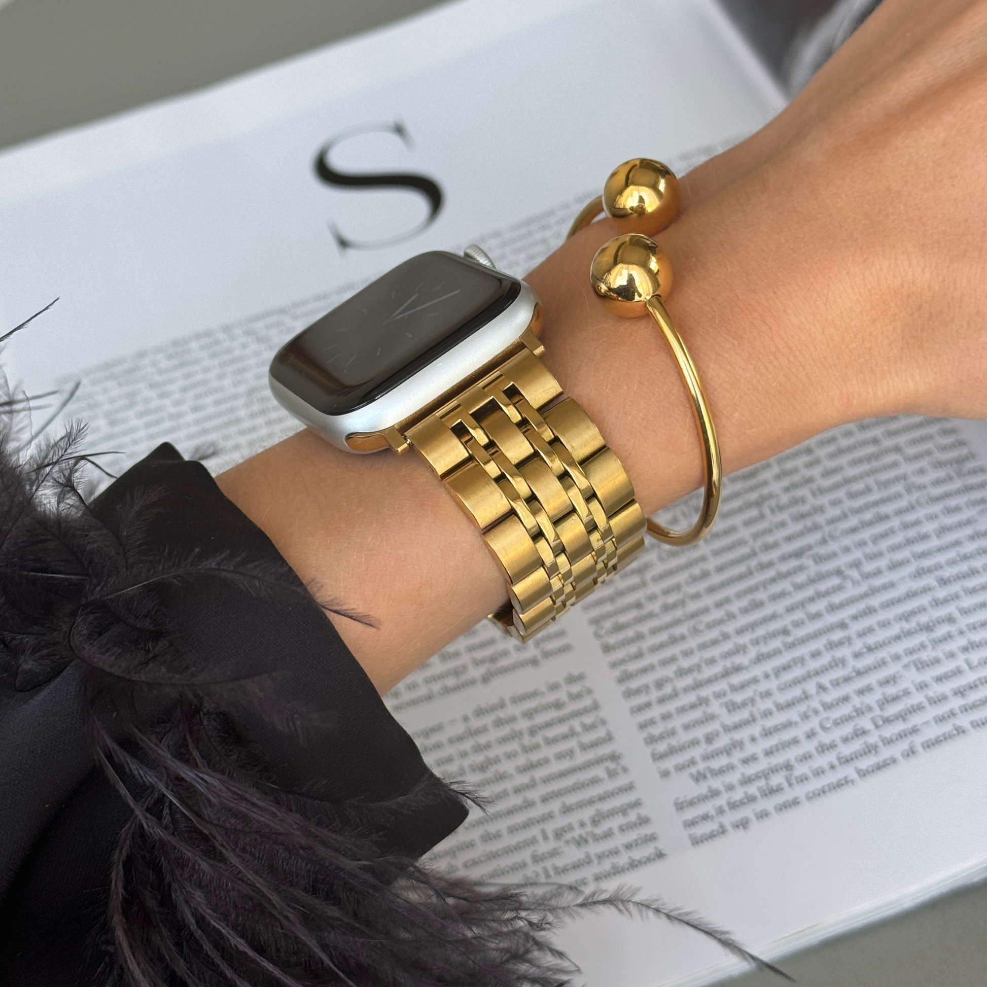 Gold link Apple Watch band worn with a gold bangle beside an open magazine. Classy stainless steel iWatch bracelet with a secure clasp, compatible with Apple Watch 38mm to 49mm, Series 1-11, SE and Ultra
